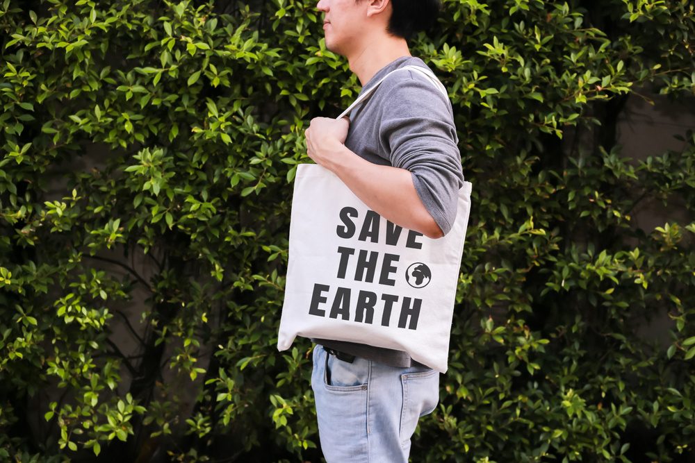Young male hipster holding and carrying the Save The Earth tote handbag in green nature environment background - ecology and recycle concept.